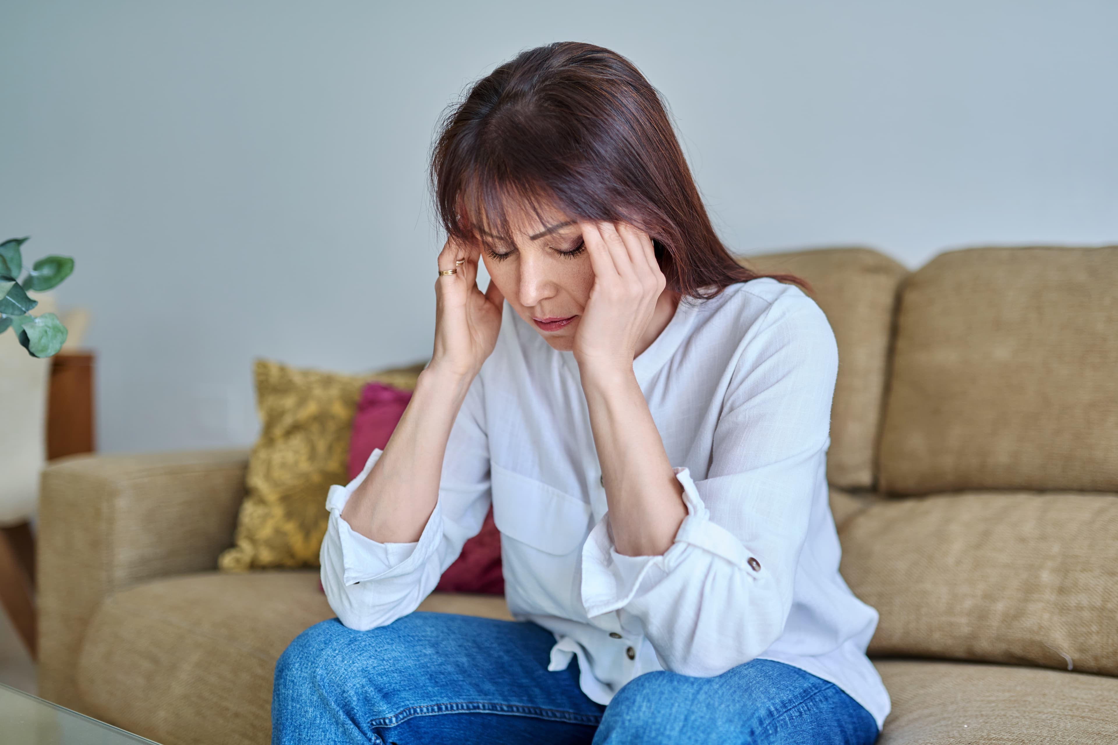 Mulher de meia-idade sentada no sofá com as mãos na cabeça e olhos fechados, expressando cansaço emocional e sobrecarga mental.